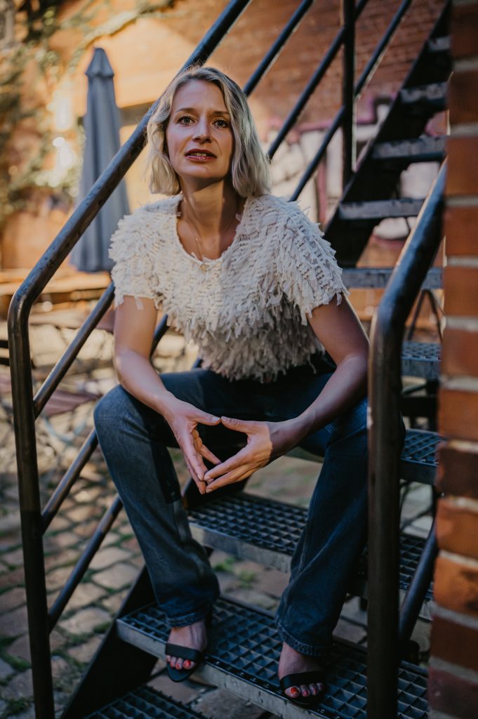 Annika Brönstrup sitzt auf einer Treppe, Blick direkt, offen und wach, Mund leicht geöffnet; Hände zu Raute geformt